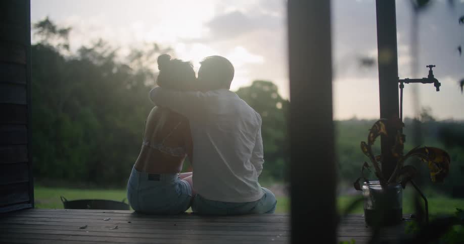 Young couple sitting on the porch looks at the sunset while hugging and kissing