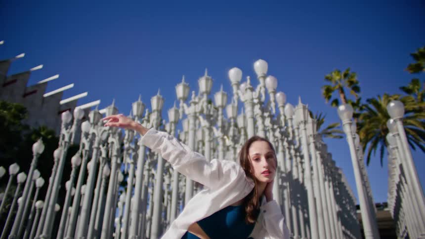 Woman in a blue top and white shirt stretches her arm forward as she dances near the light posts and palm trees under a blue sky