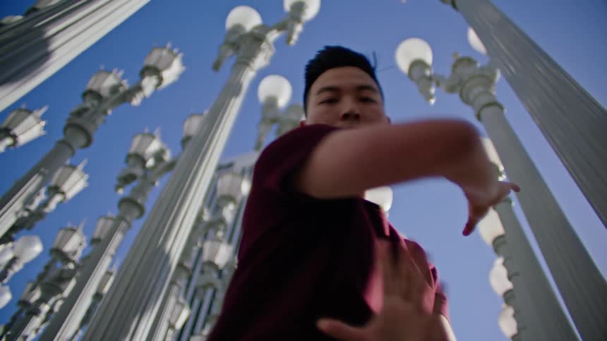 Young asian man energetically dancing in front of a streetlamps on a park under a blue sky