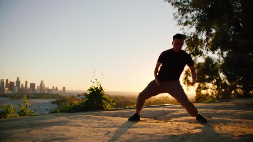 Dancing man wearing black t shirt and brown trousers is silhouetted against city skyline and sunset