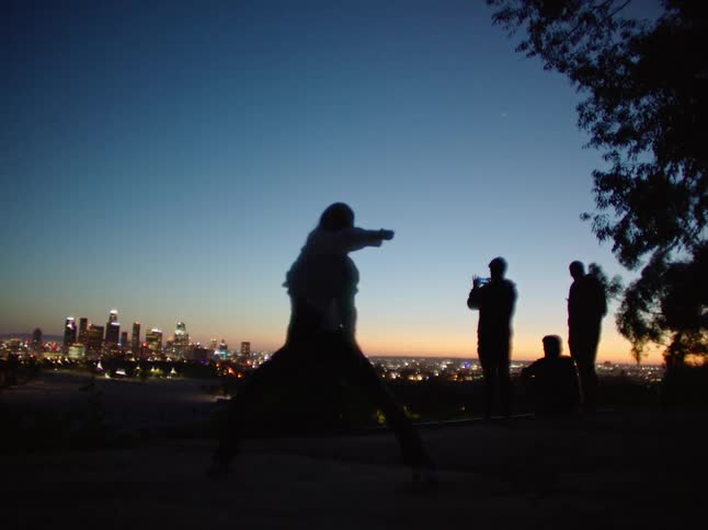 Womans silhouette dances as others stand and make video with city in background at night