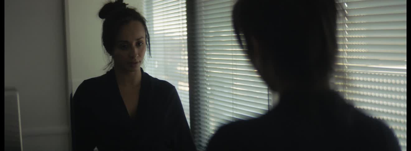 Woman with hair in bun and black jacket looks in mirror next to window blinds