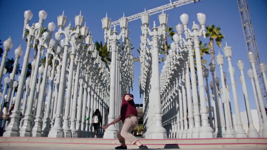 Man wearing a red shirt dances as two women walk between rows of lamp posts under a blue sky