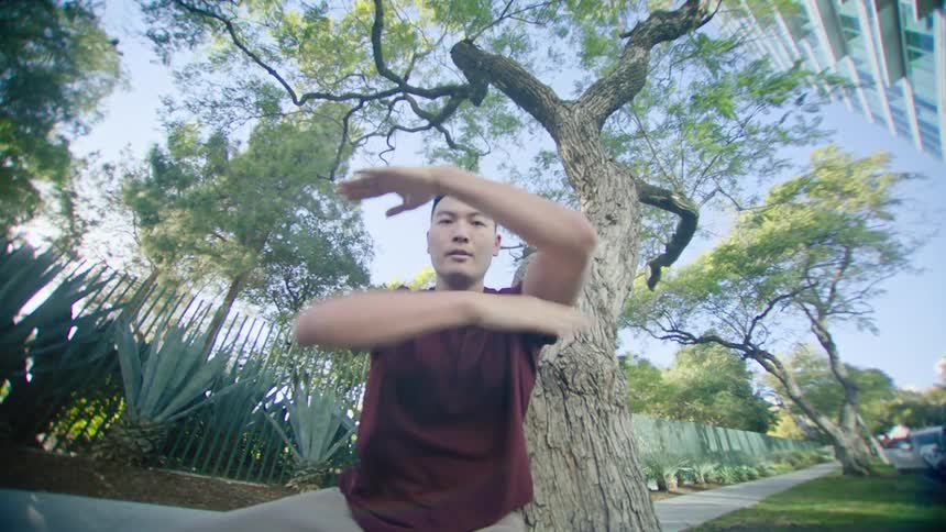Man with red t shirt and brown pants dances next to tree in the city street