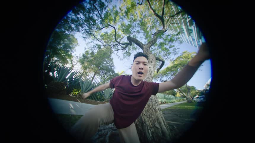 Young asian man energetically dancing in front of a tree in a park at morning through a fish eye lens