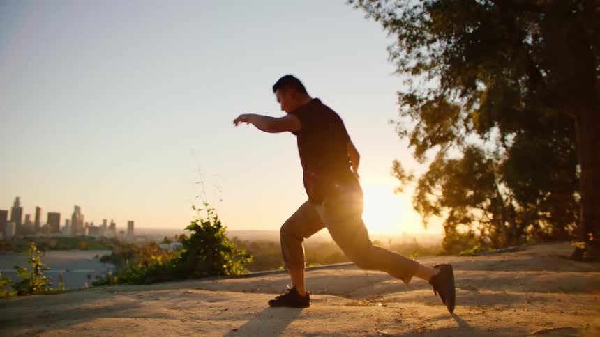 Male dancer with red shirt and brown pants dances on hill and is being silhouetted against the sunset next to city skyline