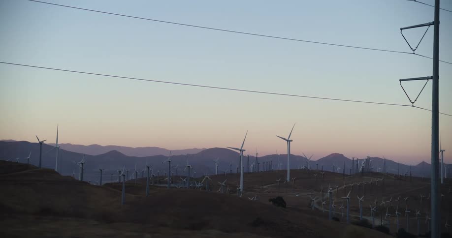 Many wind turbines are spinning on a wind farm under a clear sky with mountains in the distance