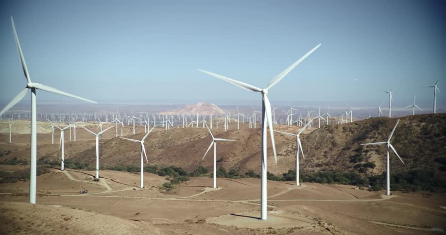 Many wind turbines are spinning on a wind farm under a clear sky with mountains in the distance