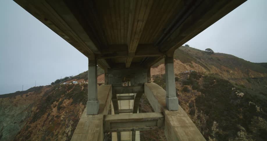 An automobile travels over a bridge high above the water as waves crash into rocks below