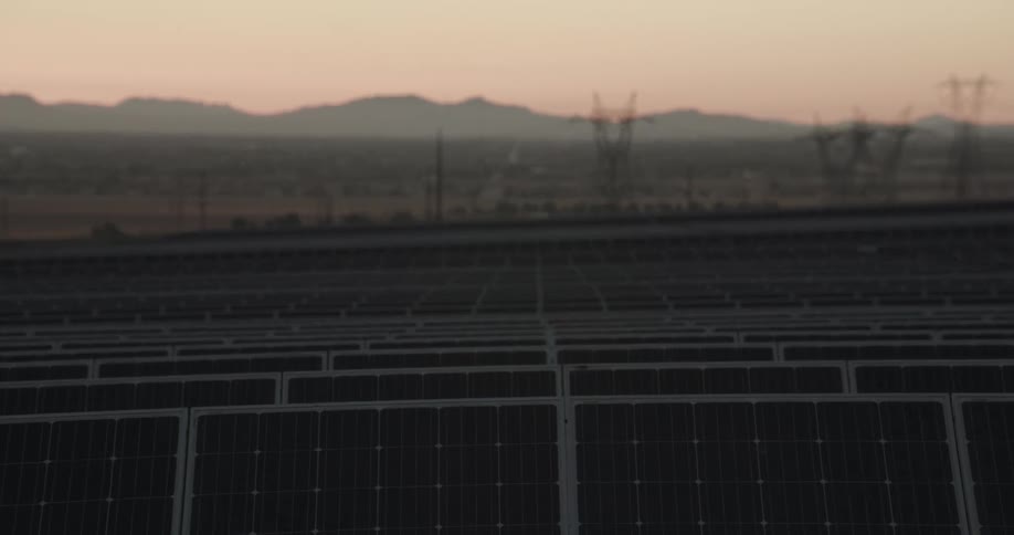 Rows of solar panels line up in a large field outdoors near transmission towers close to the mountains at sunset