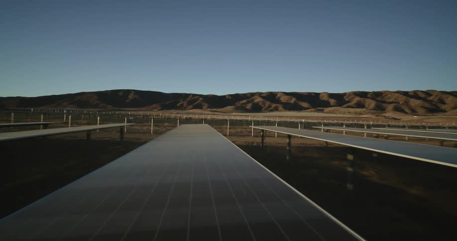 Solar panels on a solar farm reflect the shining sun on the horizon and there are mountains in the distance
