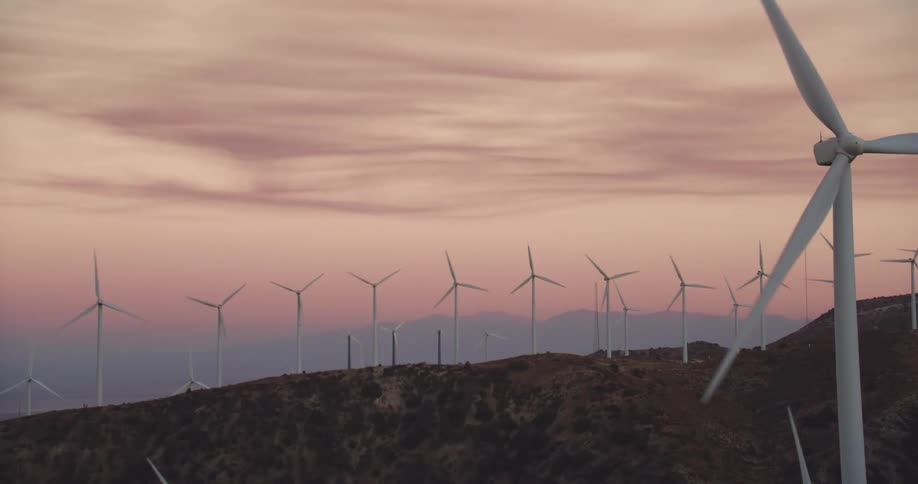 Wind turbines are spinning on a wind farm along the top of a mountain ridge under a pink sky