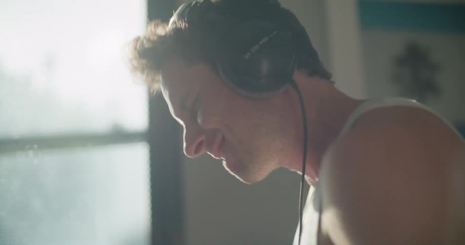 Man with white shirt and black headphones plays drums as the sun shines through windows