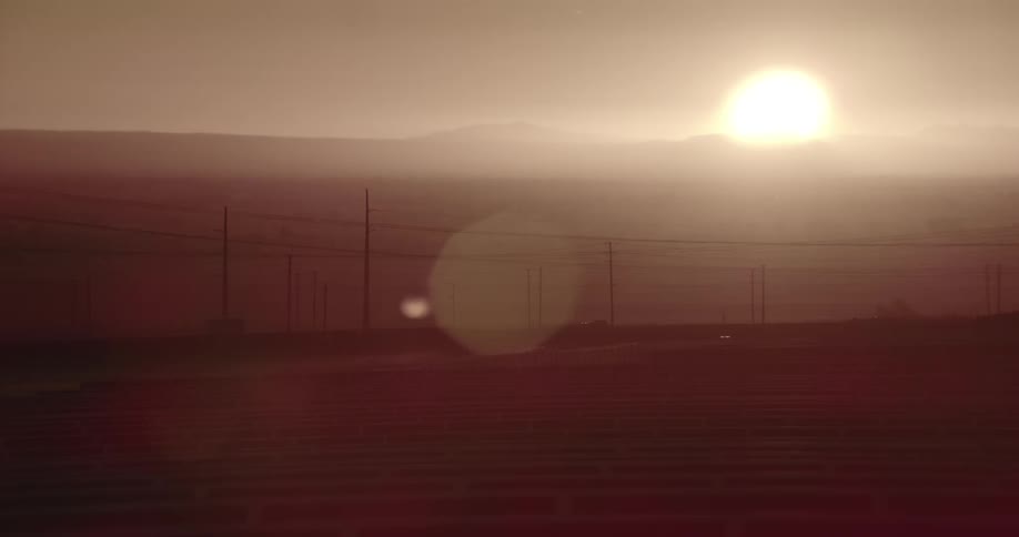 Silhouettes of utility poles and power lines as the sun shines brightly over mountains on the horizon