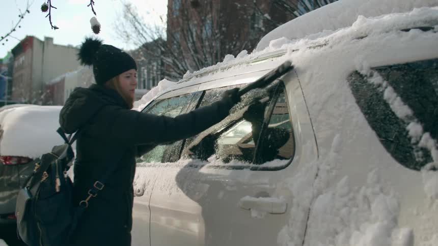 Woman wearing winter clothing using an ice scraper to take the snow out of a white cars window parked at the street