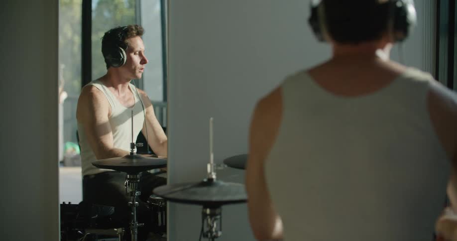 Man wearing headphones playing drums in front of a mirror next to a big window