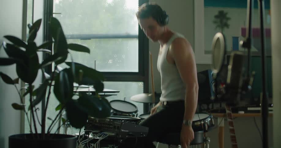 Man wearing headphones playing drums indoors next to a glass window and then leaving frustrated