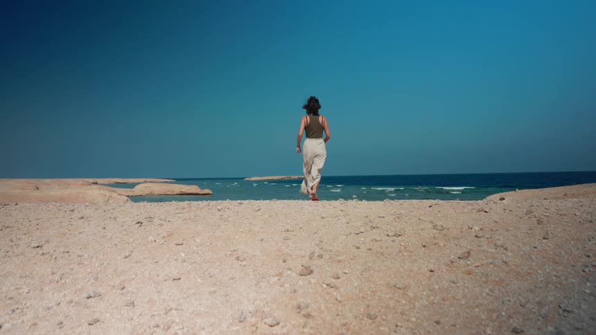 Woman with wavy hair green top beige pants and red flip flops walks to sea on the beach