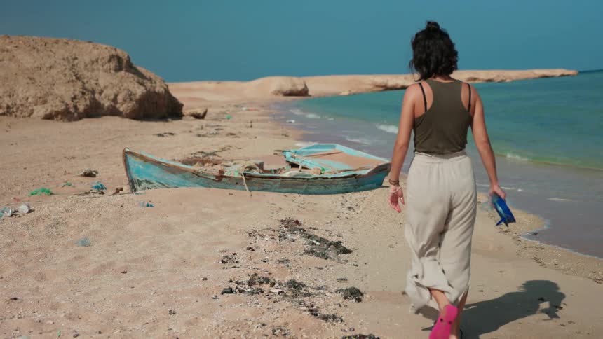 Woman walking towards a blue ruined rowboat on the sand of a beach next to the ocean