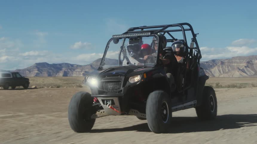 Four people in a dune buggy on a dirt road in the desert with mountains in the distance and a truck and trailer nearby