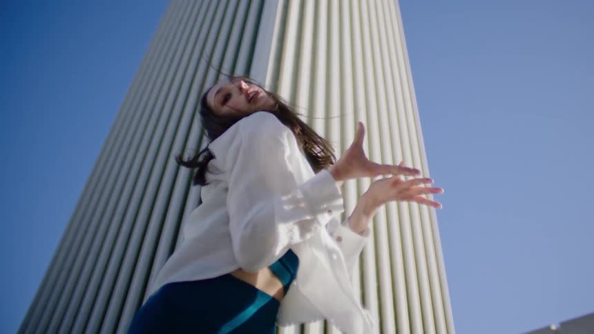 A female dancer performs a series of dance moves outside in front of a tall white building