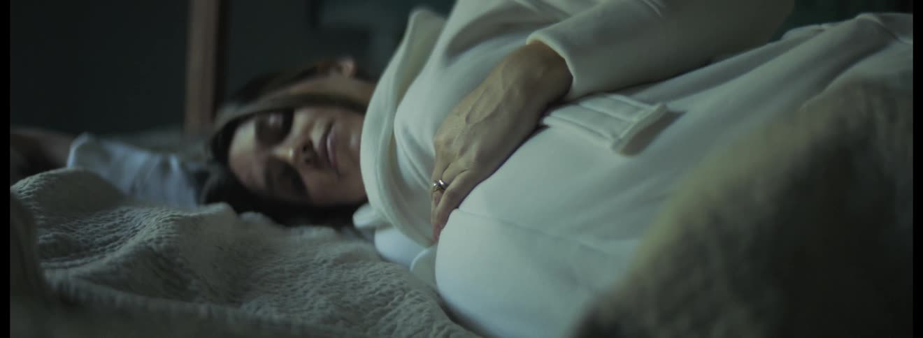 Pregnant woman with dark hair white dress and ring strokes her belly affectionately lying down on bed
