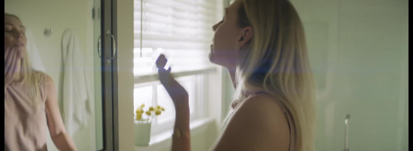 Young blonde woman enters the bathroom and takes a pill with a glass of water before looking in the mirror