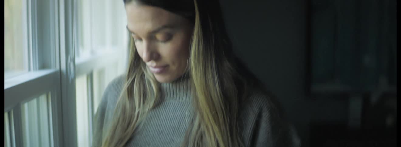 White woman wearing a sweater standing next to a big window looking outside and running her hand through her pregnant belly