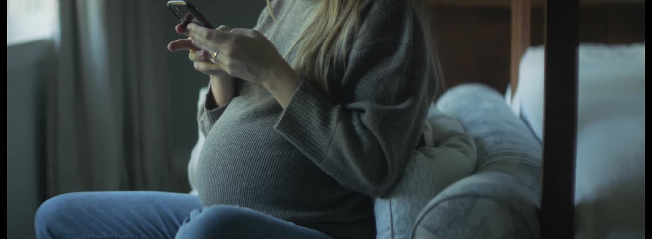 Blonde pregnant woman with gray sweater and jeans sits on couch touching her belly and using smartphone