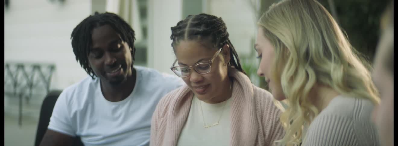 Blonde woman hands a card to a black woman who looks at it and smiles as her husband strokes her arm affectionately