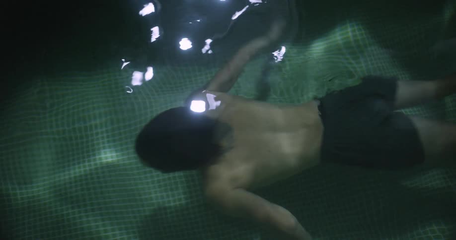 Young man wearing shorts swims underwater on an indoors pool with green tiles