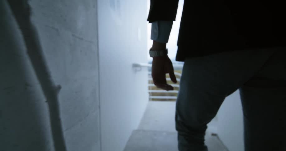 Man wearing formal clothes and a wristwatch walking through a hallway towards the stairs of a building
