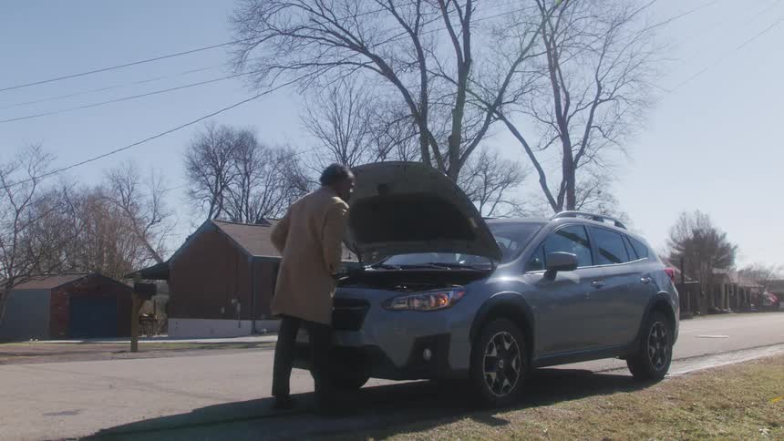 Man exits a car parked on the curb of a road to open the hood and make a phone call