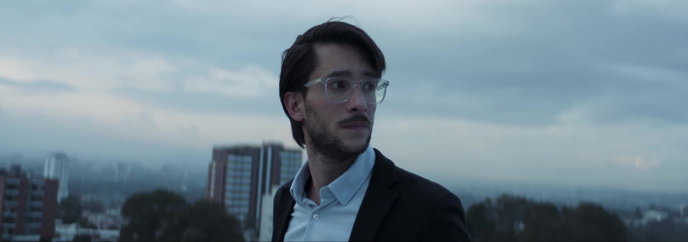 Man wearing glasses and formal clothes standing in the top of a building running his hand through his hair and looking around a big city