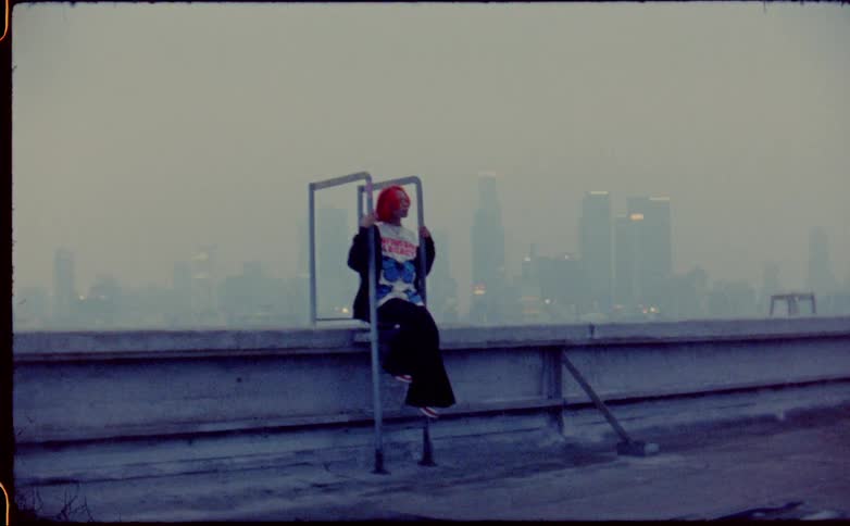 Woman with brightly dyed red hair sits on wall near ladder as fog obscures city skyline in the background