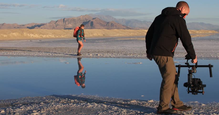 Man photographing a woman wearing backpack walking next to water