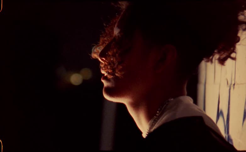A teenage boy leaning against a building outside at night adjusts his shirt