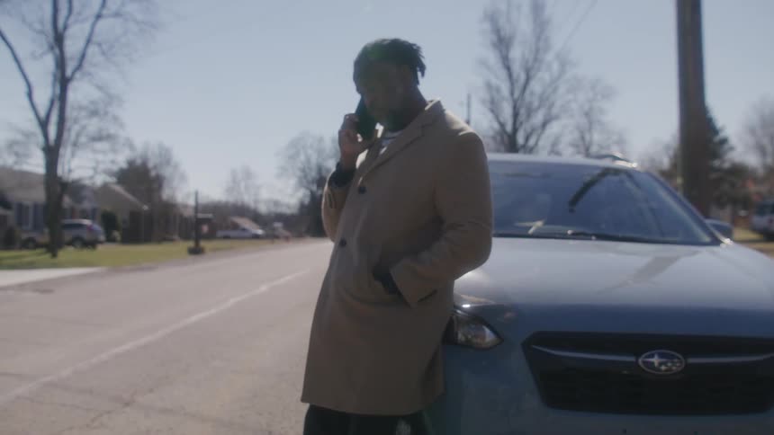 A man holds his smartphone to his ear while leaning against a car parked on a road in a residential neighborhood during the day