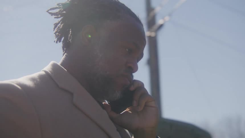 Man with dreadlocks beard and brown overcoat calls someone with smartphone in the street