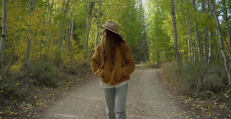 Woman with brown hat straight long hair coat and jeans walks down forest road in the woods