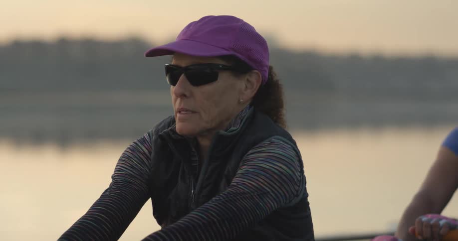 Woman rows with partner in boat along water reflecting woods on opposite shore