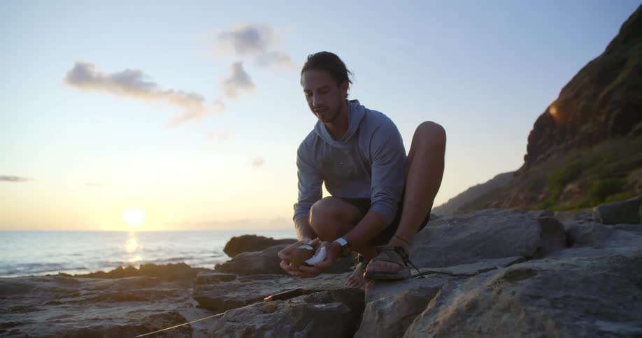 Crouching man smiles as he slices and eats piece of coconut as ocean in background reflects light from sun sitting on the horizon