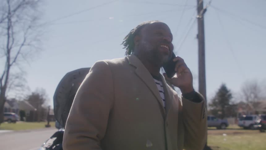 Black man wearing a jacket talks on a cell phone as he walks behind a vehicle in a neighborhood under a blue sky