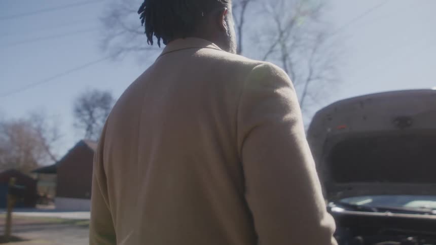 Man with dreadlocks and beard puts smartphone in his brown overcoat pocket and walks towards car in the street
