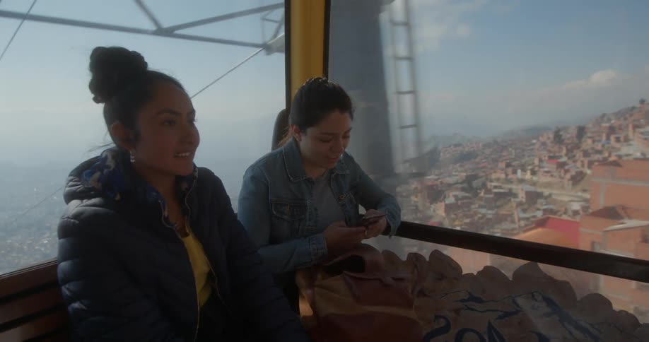 Two teenaged girls travel up the mountain in a cable car as one girl looks at her cell phone and the other smiles and nods her head