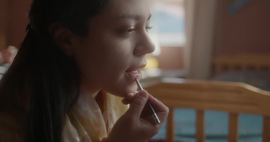 Woman applies lipstick to her lips using a lipstick pencil as she sits at a table containing makeup and bottles