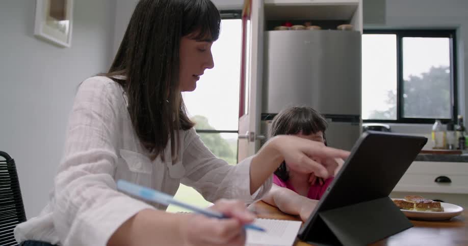Mother helps daughter with homework as they consult tablet screen