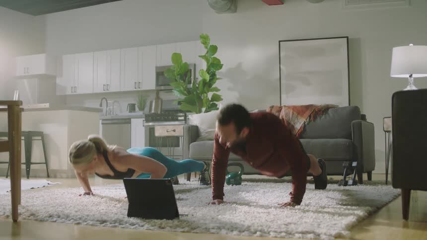 Man and woman are exercising in a living room and watching a laptop