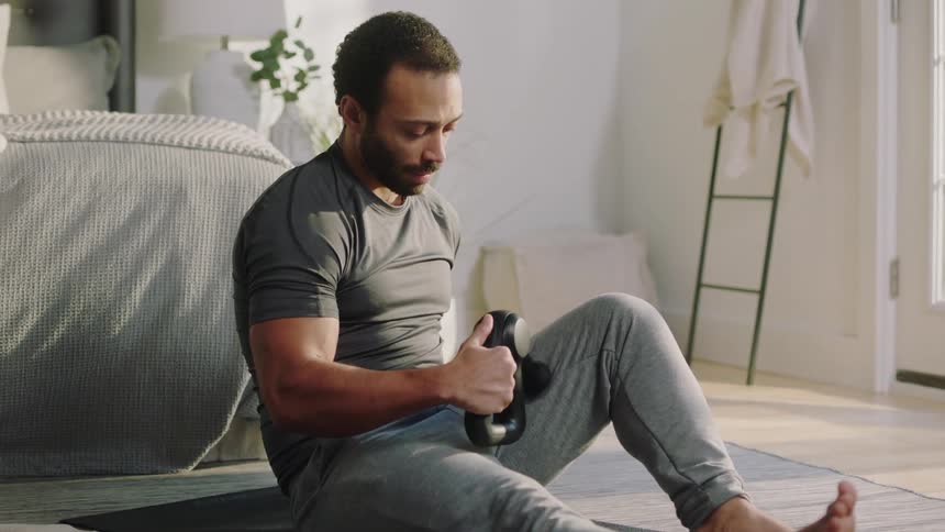 A man sitting on his bedroom floor massaging his leg muscles using a hand held power massager