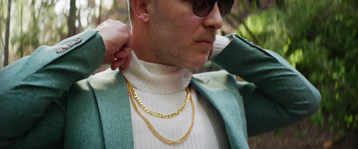 A man in gold chains adjusting his white turtleneck outdoors